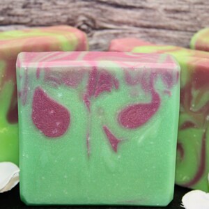 May include: Close-up of several handmade soap bars. The soap has a green base color with swirled patterns of pink and purple. The bars are rectangular and have a smooth texture. White flowers are in the foreground.