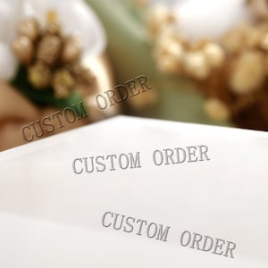 May include: A white surface with the words "CUSTOM ORDER" printed in a gray font. The text is repeated multiple times across the surface. The background is blurred, featuring white and gold floral elements.