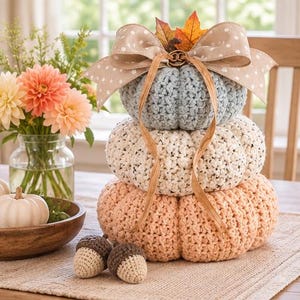 May include: A stacked crochet pumpkin decoration featuring three tiers in light blue, cream, and peach. The top is adorned with a beige polka-dot ribbon, a brown twine bow, and artificial autumn leaves. Two small crochet acorns sit at the base.