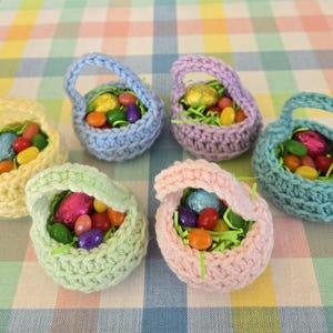 May include: Six small, crocheted Easter baskets in pastel colors: yellow, blue, purple, green, pink, and teal. Each basket is filled with colorful jelly beans, foil-wrapped chocolate eggs, and green paper grass, set on a checkered tablecloth.