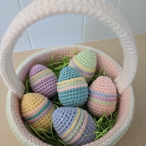 May include: A crocheted Easter basket filled with six colorful, striped crochet eggs. The basket is white with pastel green and pink accents, and a white handle. The eggs are in shades of yellow, blue, purple, and pink.