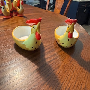 May include: Two ceramic chicken-shaped bowls with yellow bodies, red combs, and white interiors. The bowls are on a wooden table, with other chicken figurines in the background. The bowls are decorative and functional.