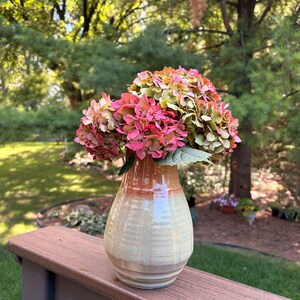 May include: A ceramic vase with a glossy finish featuring a gradient of cream and tan colors at the base transitioning to a warm orange hue at the neck. The vase contains a cluster of hydrangea flowers with petals in shades of pink, green, and brown.