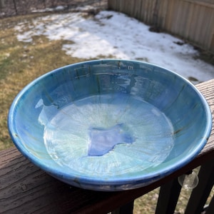May include: A handmade ceramic bowl with a glossy, multi-toned blue glaze. The bowl's interior features shades of blue and green, creating a water-like effect. The bowl is sitting on a wooden deck.