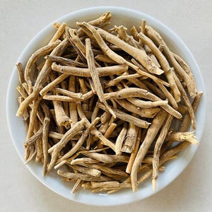 Puede incluir: Un cuenco de cer&aacute;mica blanco lleno de ra&iacute;ces de ashwagandha secas de color marr&oacute;n claro. Las ra&iacute;ces son de varios tama&ntilde;os y formas, con una apariencia &aacute;spera y texturizada. El cuenco est&aacute; sobre un fondo liso y claro.