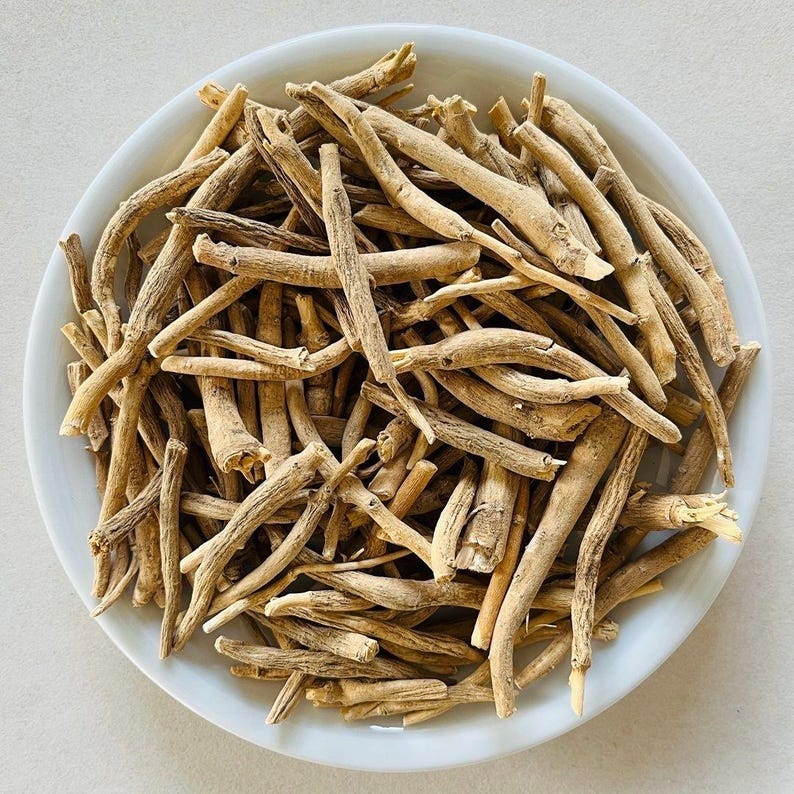 Puede incluir: Un cuenco de cer&aacute;mica blanco lleno de ra&iacute;ces de ashwagandha secas de color marr&oacute;n claro. Las ra&iacute;ces son de varios tama&ntilde;os y formas, con una apariencia &aacute;spera y texturizada. El cuenco est&aacute; sobre un fondo liso y claro.