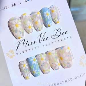 Könnte beinhalten: Ein Set mit 10 falschen Nägeln mit einem weißen und blauen Gänseblümchen-Design. Die Nägel sind klar und haben eine glänzende Oberfläche. Die Nägel sind mit "Miss Vee Bee Handmade Adornments" beschriftet.