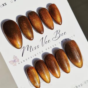 Könnte beinhalten: Ein Set handbemalter Press-On-Nägel in einem warmen Braun- und Goldfarbschema. Die Nägel haben eine ovale Form und ein glänzendes Finish. Der Text "Miss Vee Bee" und "HANDPAINTED PRESS-ON NAILS" ist sichtbar.