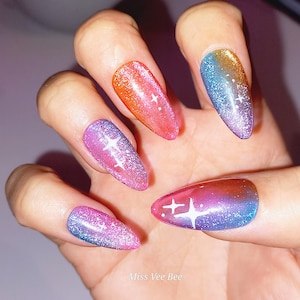 Könnte beinhalten: Nahaufnahme einer Hand mit fünf langen, mandelförmigen Nägeln, die mit einem Farbverlauf von Rosa, Orange, Blau und Lila lackiert sind. Jeder Nagel hat ein weißes Sternendesign.