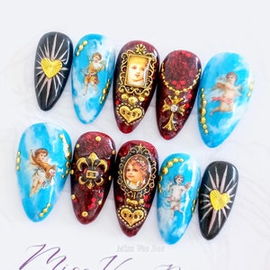 Puede incluir: Conjunto de diez uñas postizas con varios diseños. Los diseños incluyen querubines, corazones, una flor de lis y una cruz. Las uñas están pintadas en tonos de rojo, azul y negro. El texto "Baby" aparece en dos de las uñas.