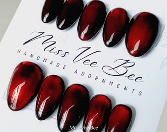 May include: Set of ten red and black oval-shaped fake nails with a marbled effect. The nails are arranged in two rows on a white surface. The text "Miss Vee Bee Handmade Adornments" is printed on the surface.