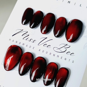 Pode incluir: Conjunto de dez unhas postiças ovais de cor vermelha e preta com efeito marmorizado. As unhas estão dispostas em duas filas sobre uma superfície branca. O texto "Miss Vee Bee Handmade Adornments" está impresso na superfície.