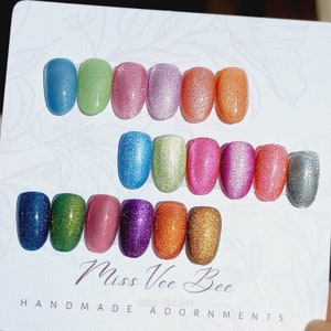 May include: A set of 14 small nail polish bottles with a variety of colors, including blue, green, pink, purple, orange, and silver. The bottles are arranged in two rows on a white background. The text "Miss Vee Bee Handmade Adornments" is printed at the bottom of the image.