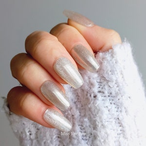 Könnte beinhalten: Nahaufnahme einer Hand mit langen, weißen, glitzernden Nägeln in Sargform. Die Nägel sind mit einem schimmernden, irisierenden Finish lackiert.