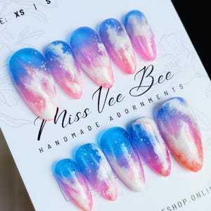 Könnte beinhalten: Set mit zehn falschen Nägeln mit einem blauen und rosa Ombre-Design und weißen Wolkenakzenten. Die Nägel sind in eine lange ovale Form gebracht und haben eine glänzende Oberfläche. Der Text "Miss Vee Bee HANDMADE ADORNMENTS" ist auf dem weißen Hintergrund gedruckt.