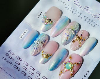 May include: A set of ten press-on nails featuring a pastel color palette of pink, blue, and white with a pearlescent finish. The nails are decorated with gold starfish charms, small white pearls, and iridescent rhinestones. The display card includes the text Miss Vee Beauty, Freshly Baked Handpainted Press-On Nails, and size, shape, and length selection boxes.