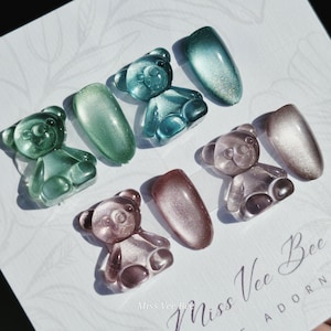 Könnte beinhalten: Eine Auswahl an Nagelkunst mit bärenförmigen Charms in Grün, Blau und Rosa, zusammen mit ovalen Nageldesigns. Die Nagelkunst wird auf einer weißen Oberfläche mit dem Text "Miss Vee Bee" präsentiert.