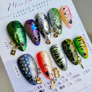 May include: A collection of hand-painted press-on nails, each designed to resemble a fishing lure. The nails feature various colours, including green, silver, and orange, with gold charms. The text "Miss Vee Bee" is visible on the card.