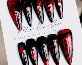 May include: Set of ten red and black stiletto press-on nails with a holographic finish.  The nails are arranged on a white card with the text "Miss Vee Bee HANDPAINTED PRESS-ON NAILS".