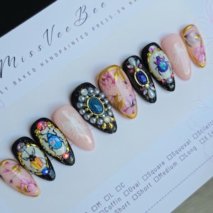 May include: A set of ten press-on nails with a black base and various designs. The designs include floral patterns, rhinestones, and a beetle motif. The nails are arranged in a row on a white surface with a size chart.