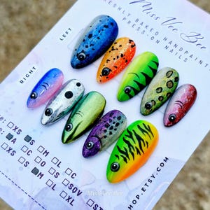 May include: A collection of ten hand-painted, almond-shaped press-on nails. The designs include blue with black spots, orange with glitter, and green with black stripes. Each nail has a fish-like design with a black eye. The text "Miss Vee Bee" is visible.