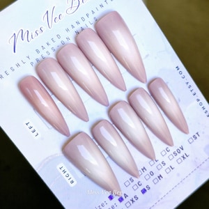 May include: Set of ten nude-colored, long, stiletto-shaped press-on nails. The nails are arranged in two rows on a white card with the text "Miss Vee Bee" and "RESHLY BAKED HANDPAINTED PRESS-ON NAILS" printed on it.