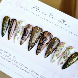 Puede incluir: Conjunto de diez uñas postizas largas en forma de estilete con varios diseños. Las uñas presentan una estética vintage con patrones florales, detalles dorados y texto que dice "RAL FLAVORS ON", "Bonne" y "BEETLEJUICE".