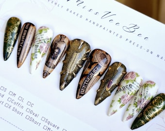 May include: Set of ten long, stiletto-shaped press-on nails with various designs. The nails feature a vintage aesthetic with floral patterns, gold accents, and text that reads "RAL FLAVORS ON", "Bonne", and "BEETLEJUICE".