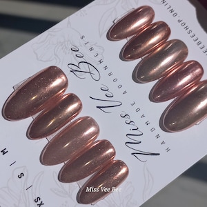 Pode incluir: Um conjunto de unhas postiças em ouro rosa com acabamento metálico. As unhas, em formato amêndoa, estão dispostas em um cartão branco com o texto "Miss Vee Bee Adornments" e motivos florais.