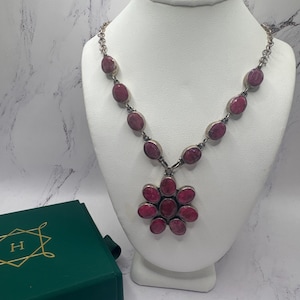 May include: A silver necklace featuring oval and flower-shaped red gemstones, displayed on a white bust. A dark green box with gold lettering is in the lower left corner. The necklace has a chain and a pendant.