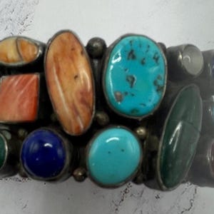 May include: A silver bracelet with a collection of colourful gemstones. The bracelet displays various shapes and colours, including turquoise, orange, blue, green, and brown stones. The stones are set in a silver metal setting.