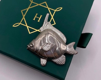 May include: A silver fish brooch on a dark green box. The brooch has detailed fins and scales. The box features a gold geometric design with the letter "H". A dark green ribbon is visible.