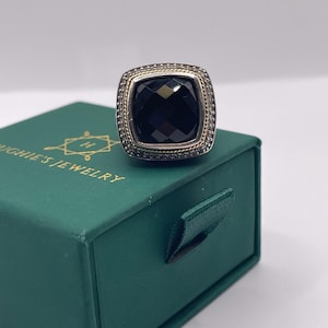 May include: A square-faced ring with a large, faceted black stone. The ring has a silver-colored band with a gold-colored rope design around the black stone. The ring is on a green jewelry box with the text "HUGHIE'S JEWELRY".