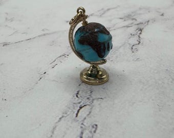 May include: A miniature globe charm with a gold-colored stand and frame. The globe features a turquoise blue ocean and brown continents. The charm is a small, detailed representation of the Earth, suitable for jewelry or craft projects.