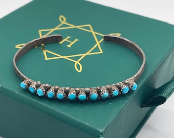 May include: A silver-toned cuff bracelet with a row of small, turquoise-colored stones. The bracelet is resting on a dark green box with a gold-colored geometric design and the letter "H".