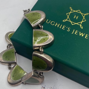 May include: A silver-toned bracelet with olive green enamel segments. The bracelet is composed of connected, half-moon shaped links. The bracelet is resting on a green box with gold text that reads "UGHIE'S JEWELRY".