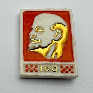 Può includere: Spilla rettangolare in ceramica con un profilo dorato di un uomo su sfondo bianco, incorniciato da un bordo arancione. Il numero "100" è in oro. Un oggetto da collezione vintage.