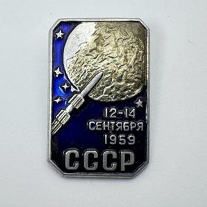 Puede incluir: Un pin rectangular vintage con borde plateado y fondo de esmalte azul. El pin presenta una luna plateada texturizada, un cohete, estrellas y texto en ruso. El texto dice "12-14 Septiembre 1959" y "CCCP".