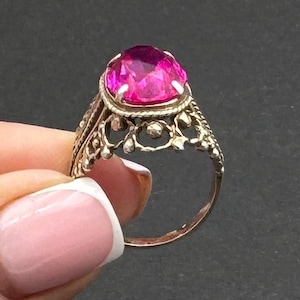 May include: A vintage gold-coloured ring with a large, oval-cut, bright pink gemstone. The ring has an ornate, filigree design around the gemstone and along the band, showcasing intricate details and a classic aesthetic.
