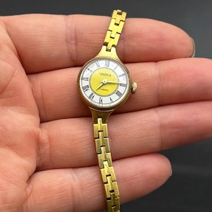 May include: A gold-tone wristwatch with a round case and a matching link bracelet. The watch face features a bright yellow center, a white outer ring with black Roman numerals, and the Cyrillic text "ЧАЙКА" printed in black above the center hands.