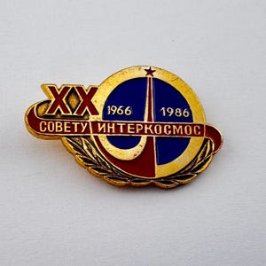 May include: Vintage Soviet-era pin with a gold-colored border and accents. The pin features a blue and red design with the text "XX" and "1966-1986" in red and white. The text "COBETУ ИHTEPKOCMOC" is also visible. The pin has a star at the top.