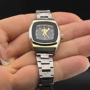 May include: A vintage wristwatch with a silver-tone metal band and a gold-tone and black watch face. The watch face has a square shape with rounded corners and features gold-tone hands and hour markers. The watch is held in a hand.