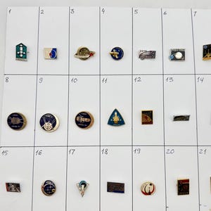 Può includere: Una collezione di ventuno spille smaltate a tema spaziale disposte in una griglia su un cartoncino bianco, numerate da uno a ventuno. Le spille presentano varie forme, tra cui cerchi, rettangoli e triangoli, con disegni di razzi, pianeti e stelle in finiture metalliche con colori blu, rosso, oro e argento.