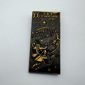 May include: A rectangular, black and gold pin celebrating the 20th anniversary of the space era. The pin features a satellite, stars, and text in Russian. The design includes a crescent moon and a star.
