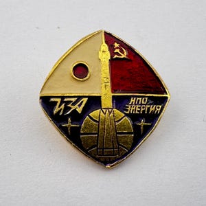 May include: Vintage Soviet-era badge with a gold-colored rocket design. The badge is divided into four sections: cream, red, blue, and gold. It features Cyrillic text and a red star and hammer and sickle symbol.