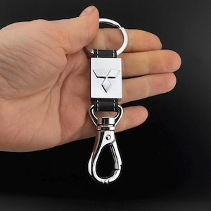 Op de afbeelding: Sleutelhanger met een zilverkleurige metalen sluiting en een zwarte leren band. De sleutelhanger heeft een zilverkleurige rechthoekige plaat met een Mitsubishi-logo. De sleutelhanger wordt in een hand gehouden.