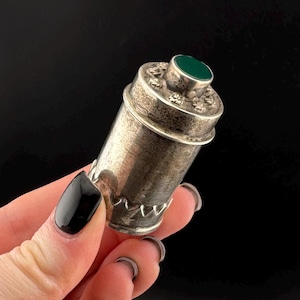May include: A silver and green metal container with a decorative design. The container has a round lid with a green stone inset.