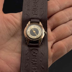 May include: Vintage wristwatch with a gold-tone case and a dark dial, set on a wide, brown leather cuff. The cuff features an embossed floral pattern. The watch has gold hands and numerals. A classic timepiece with a unique band.