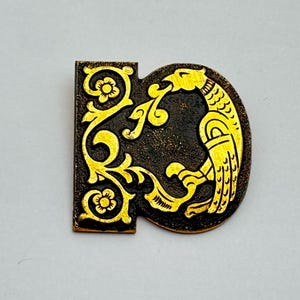 May include: A bronze-coloured metal pin featuring a gold-coloured bird design on the right and floral filigree on the left. The bird has a detailed wing and beak, and the floral design includes small flowers and swirling vines.