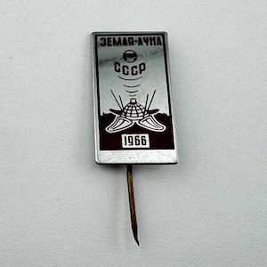 May include: A rectangular silver-colored pin with a brown border and a pin backing. The pin features the text "ЗЕМЛЯ-ЛУНА" at the top, the word "CCCP" in the center, and the number "1966" at the bottom. The pin has a design of a globe with radio waves.